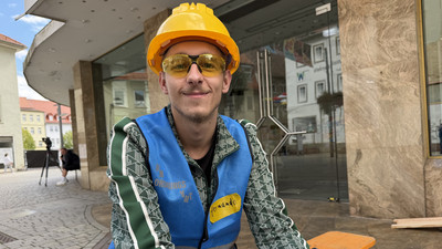Ein junger Mann in blauer Weste mit dem Namensschild "Fernando" grinst in die Kamera. Er trägt einen Bauhelm und eine Schutzbrille.