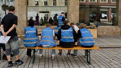 Vier Menschen mit blauen Westen mit dem Schriftzug "Umordnungsamt" sitzen auf einer Bierbank.